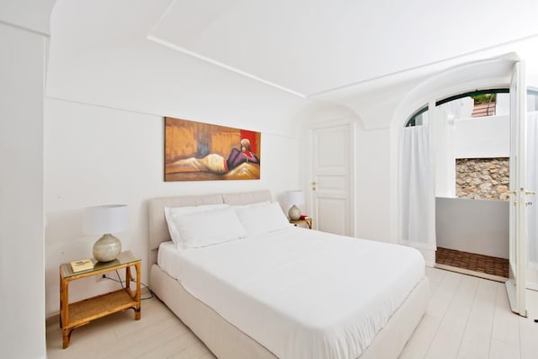 A bedroom with a large bed, two bedside tables with lamps, a painting on the wall, and an open door leading to a balcony with a stone wall.