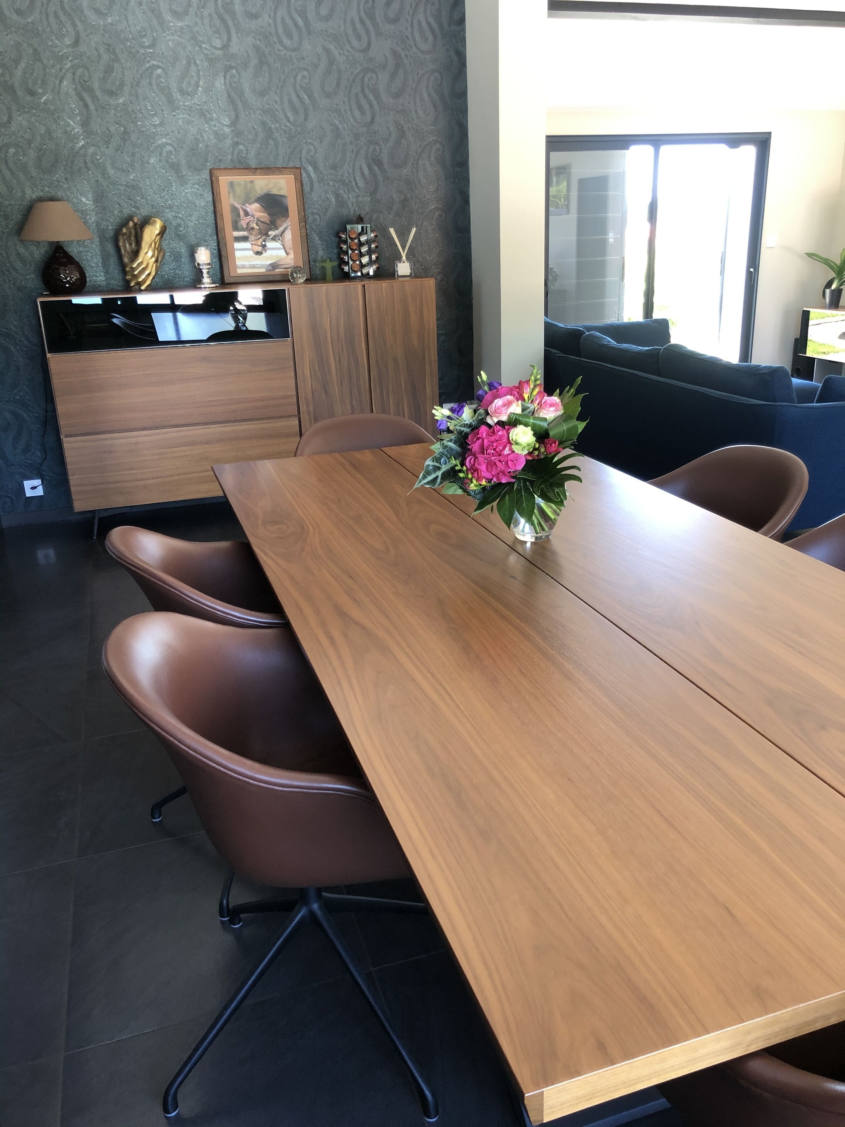 A dining area with a wooden table, brown chairs, a vase of flowers, a cabinet with a picture, and a patterned wall.