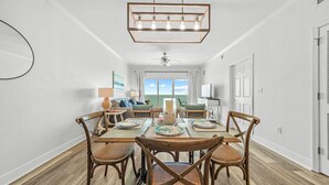 A dining area with a wooden table and chairs, a chandelier, and a view of the outdoors through a window. - Top-Rated Comfort – Guest-Praised "Nicest unit" – Gulf Front – Two King Beds (Gulf Shores)
