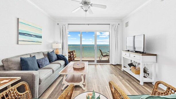 A modern living room with a sofa, coffee table, and TV stand, a ceiling fan, and a large sliding glass door leading to a balcony with a view of the ocean. - Top-Rated Comfort – Guest-Praised "Nicest unit" – Gulf Front – Two King Beds (Gulf Shores)