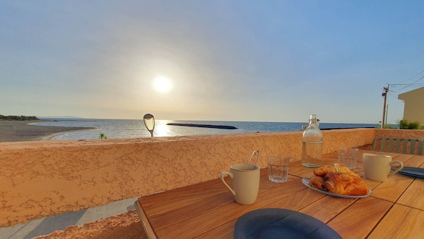 Una mesa con una botella, vasos, una taza y un plato con comida, con vista a una playa al atardecer.