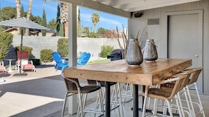 A covered patio area with a wooden table and metal chairs, surrounded by potted plants and a blue chair. - Nova Gypsy Vanner | Pool | Spacious Interior (Scottsdale)