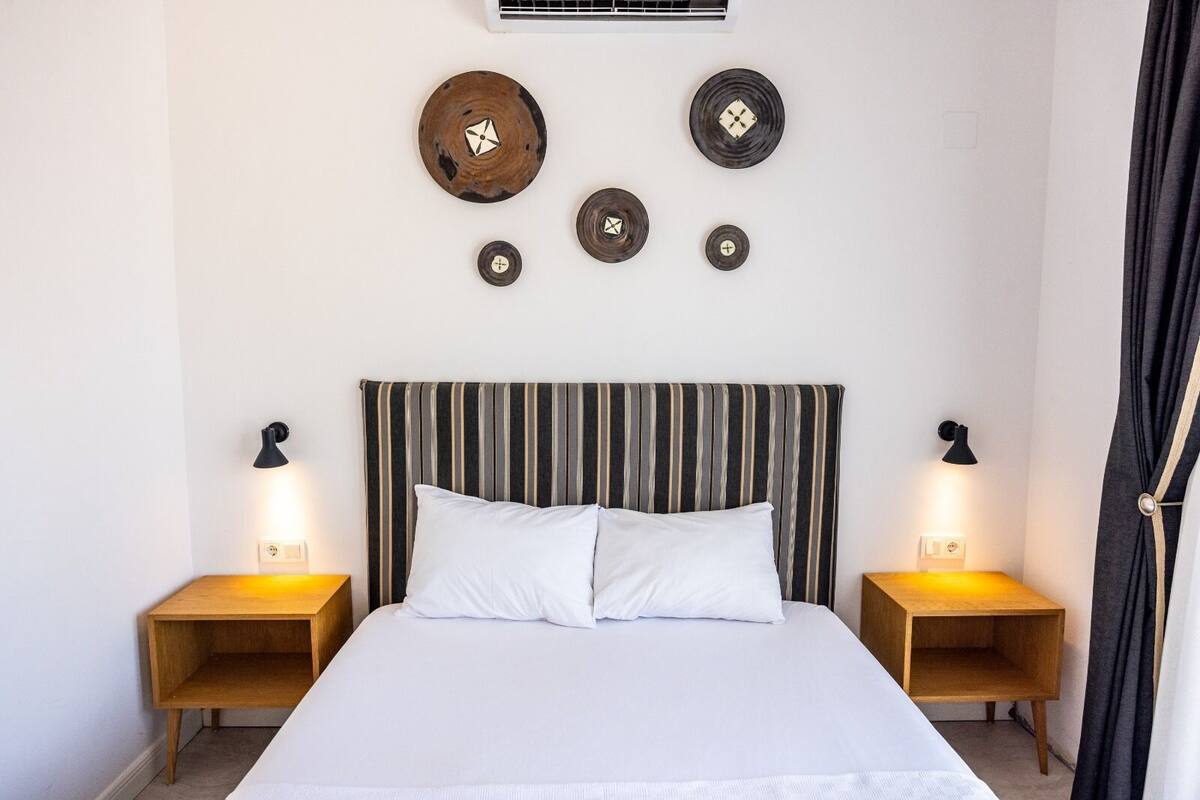 A bedroom with a striped headboard, a white bed, two bedside tables with lamps, and wall-mounted decorative plates.