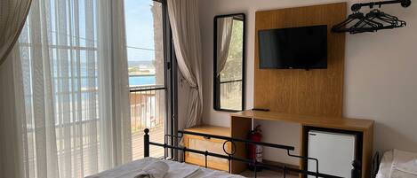 A hotel room with a bed, a TV mounted on a wooden panel, a desk with a chair, and a view of the sea through the window.