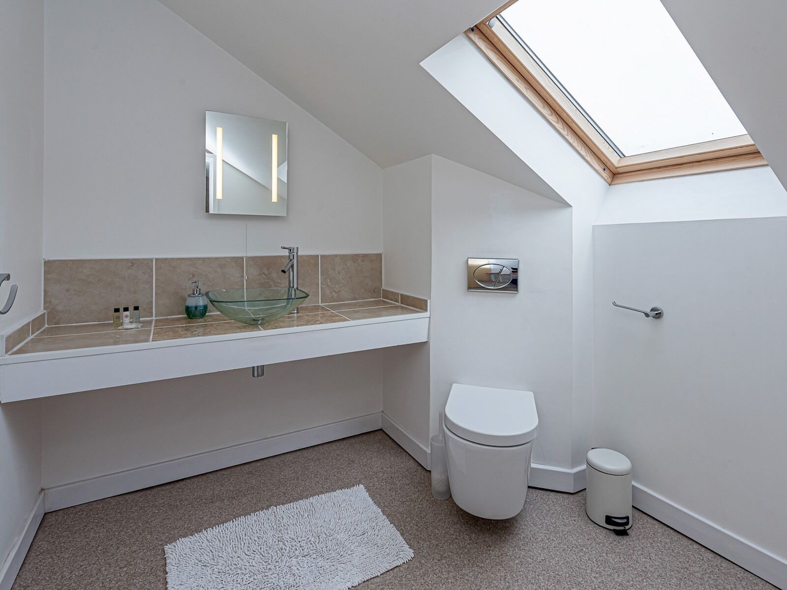 A modern bathroom with a skylight, a glass vessel sink, a toilet, and a trash bin.