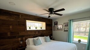 Ein Schlafzimmer mit einem großen Bett, Holzvertäfelung, einem Deckenventilator, einem Fenster mit Vorhängen und Blick ins Freie.