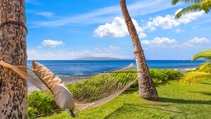 Un hamac est tendu entre deux arbres, offrant une vue imprenable sur une plage, l’océan et une île lointaine.