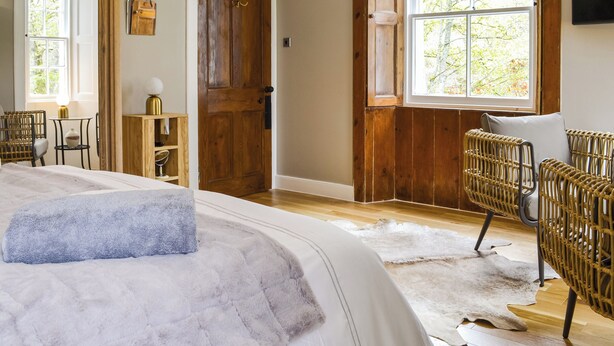 A bedroom with a large bed, a wooden door, a window with a view of trees, wicker chairs, and a small table with a lamp.
