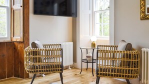 A room with two wicker chairs, a small table with a lamp, a television on the wall, and a window with a view of trees.