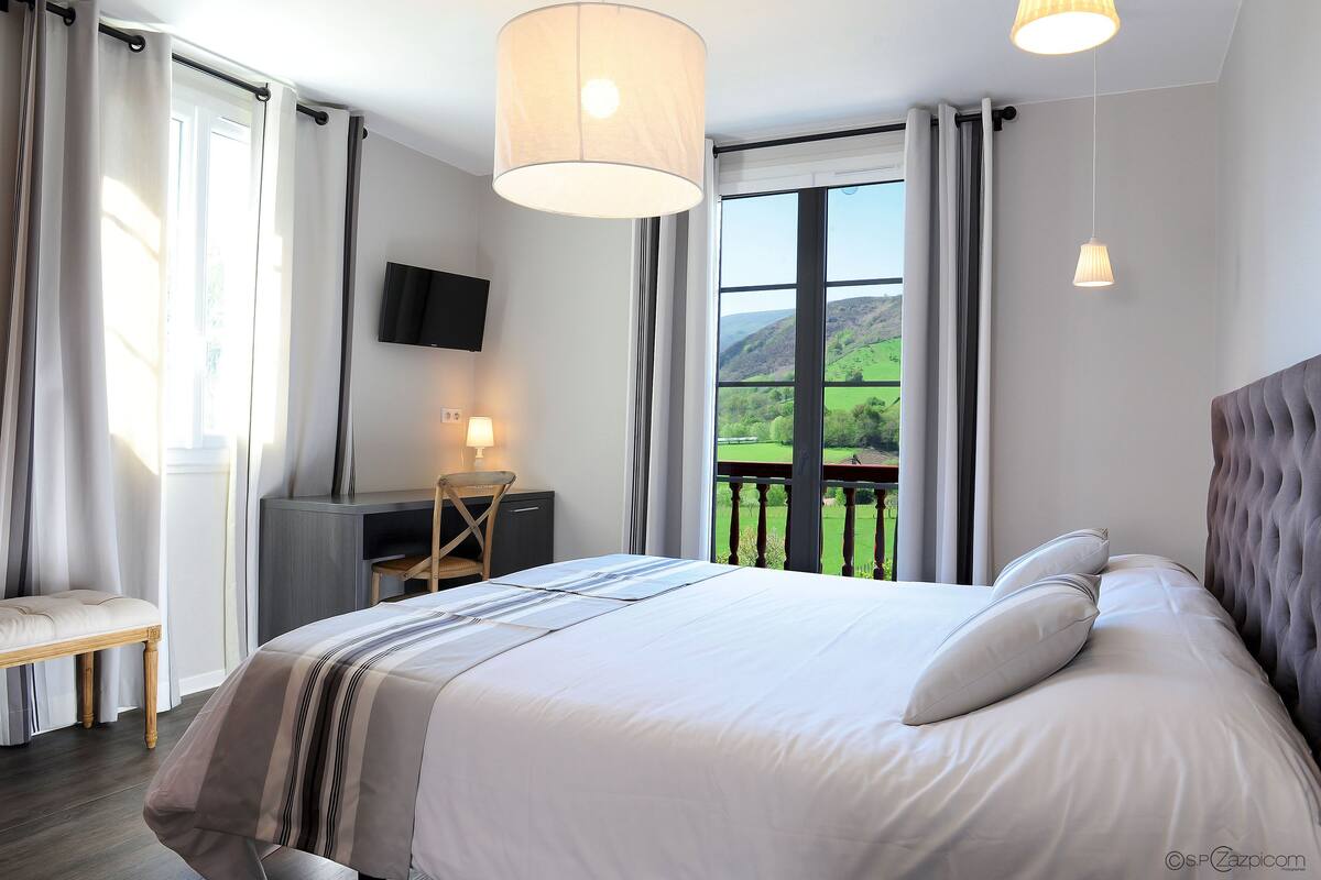A bedroom with a large bed, a desk, a chair, a TV, and a balcony with a view of the countryside.