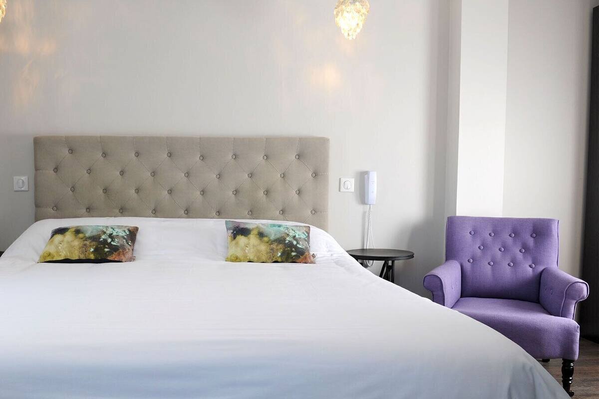 A hotel room with a large bed, two decorative pillows, a tufted headboard, a small round table, a purple armchair, and a wall-mounted phone.