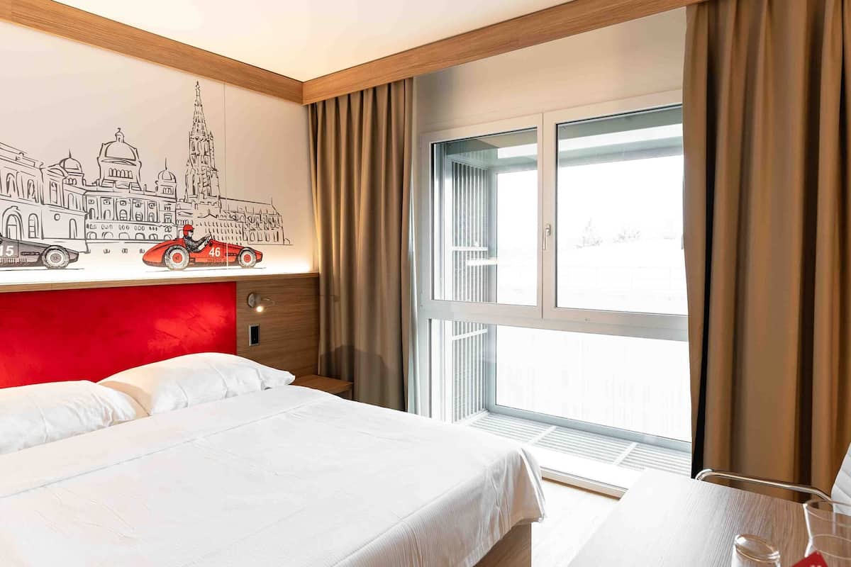 A hotel room with a large bed, a wooden desk, a window with curtains, and a wall mural.