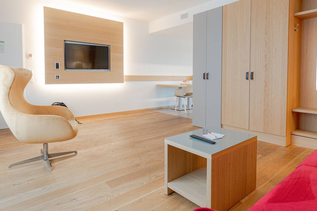 A modern living room with a wooden floor, a red sofa, a white chair, a TV mounted on the wall, and a desk with a chair.