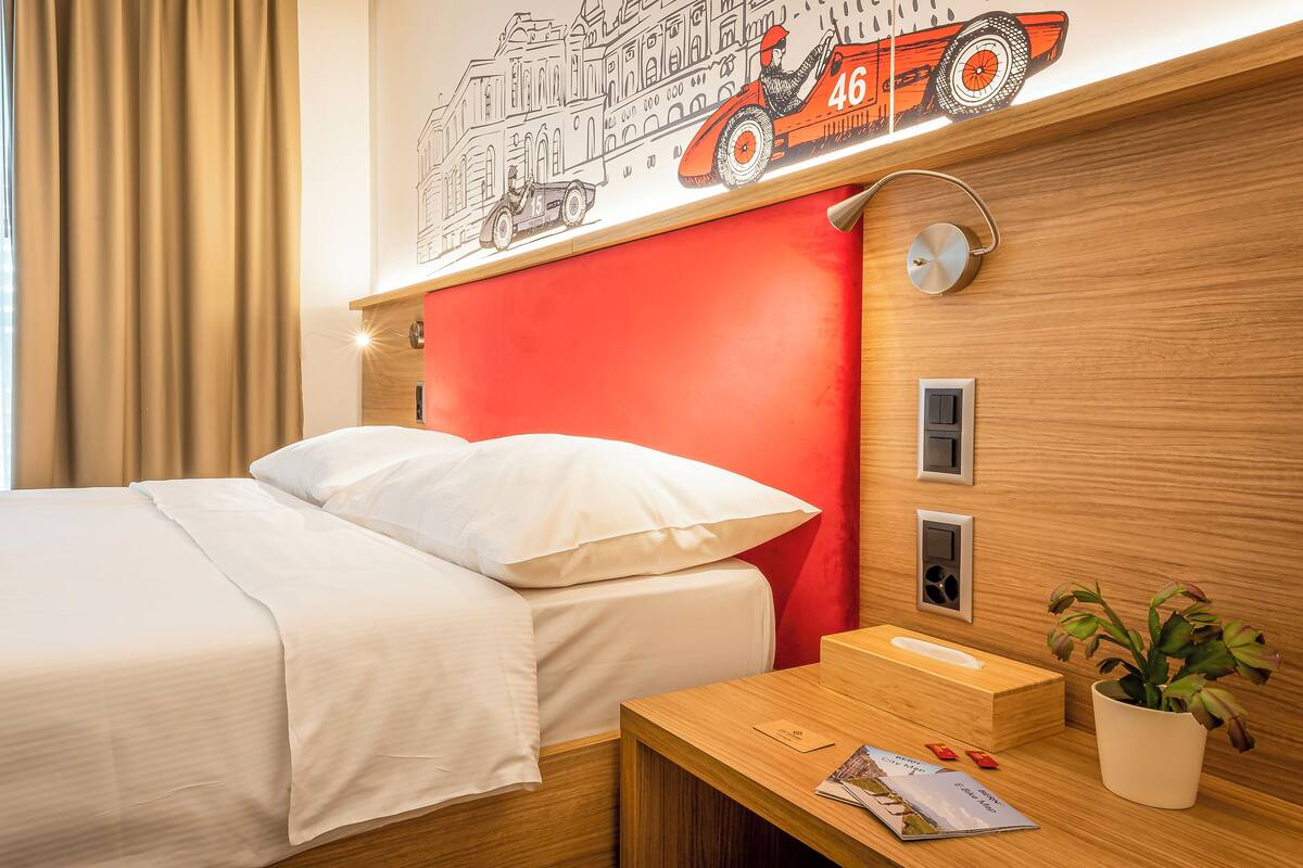 A hotel room with a bed, a nightstand, a lamp, a wall art of a racing car, and a potted plant.