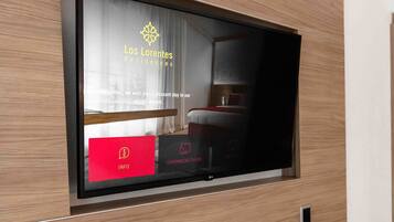 Une chambre d’hôtel moderne dotée d’un grand téléviseur à écran plat fixé au mur, d’une table de chevet en bois sur laquelle se trouve un plateau avec des verres, et d’une petite boîte en bois contenant des mouchoirs.