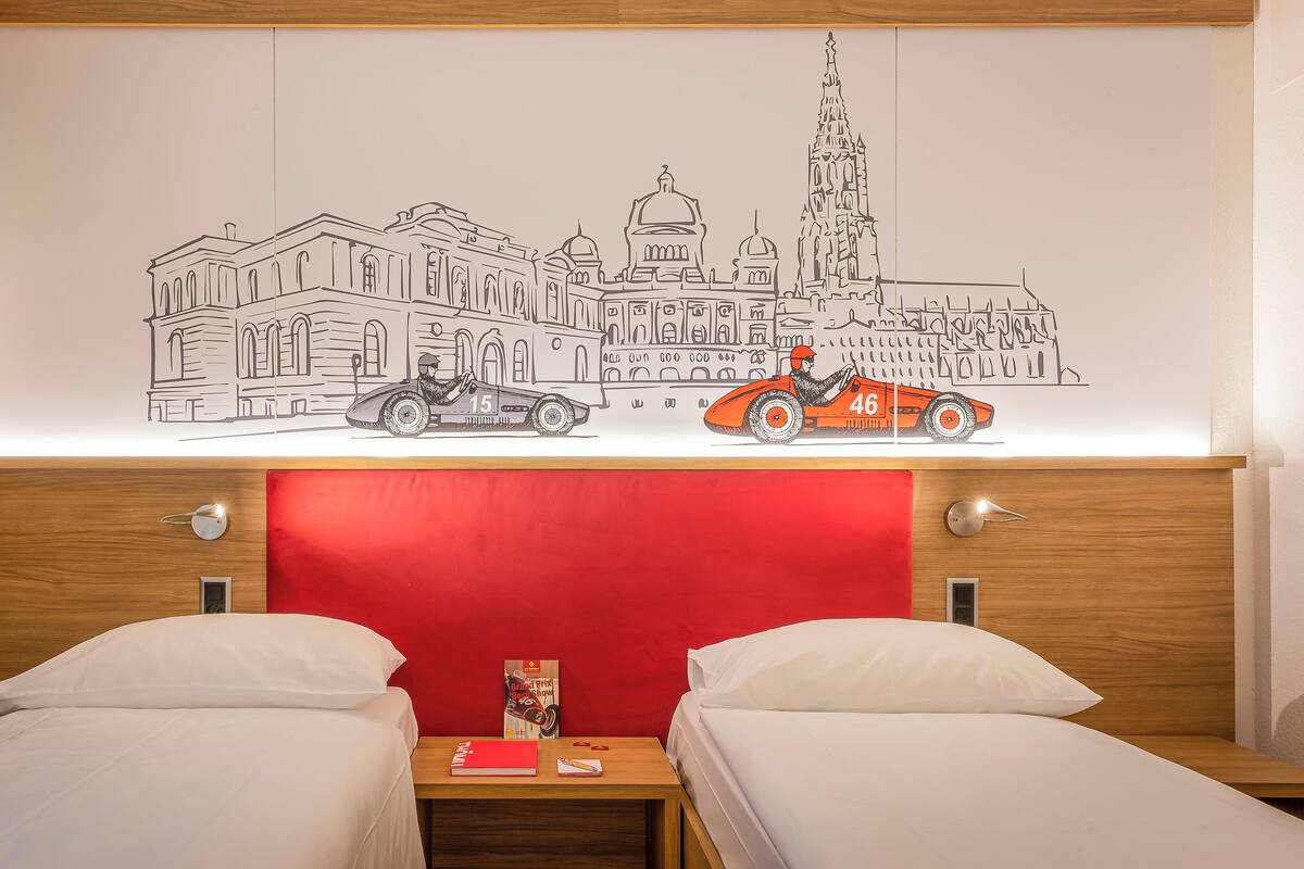 A hotel room with two beds, a red headboard, and a wall mural depicting a cityscape with cars and buildings.