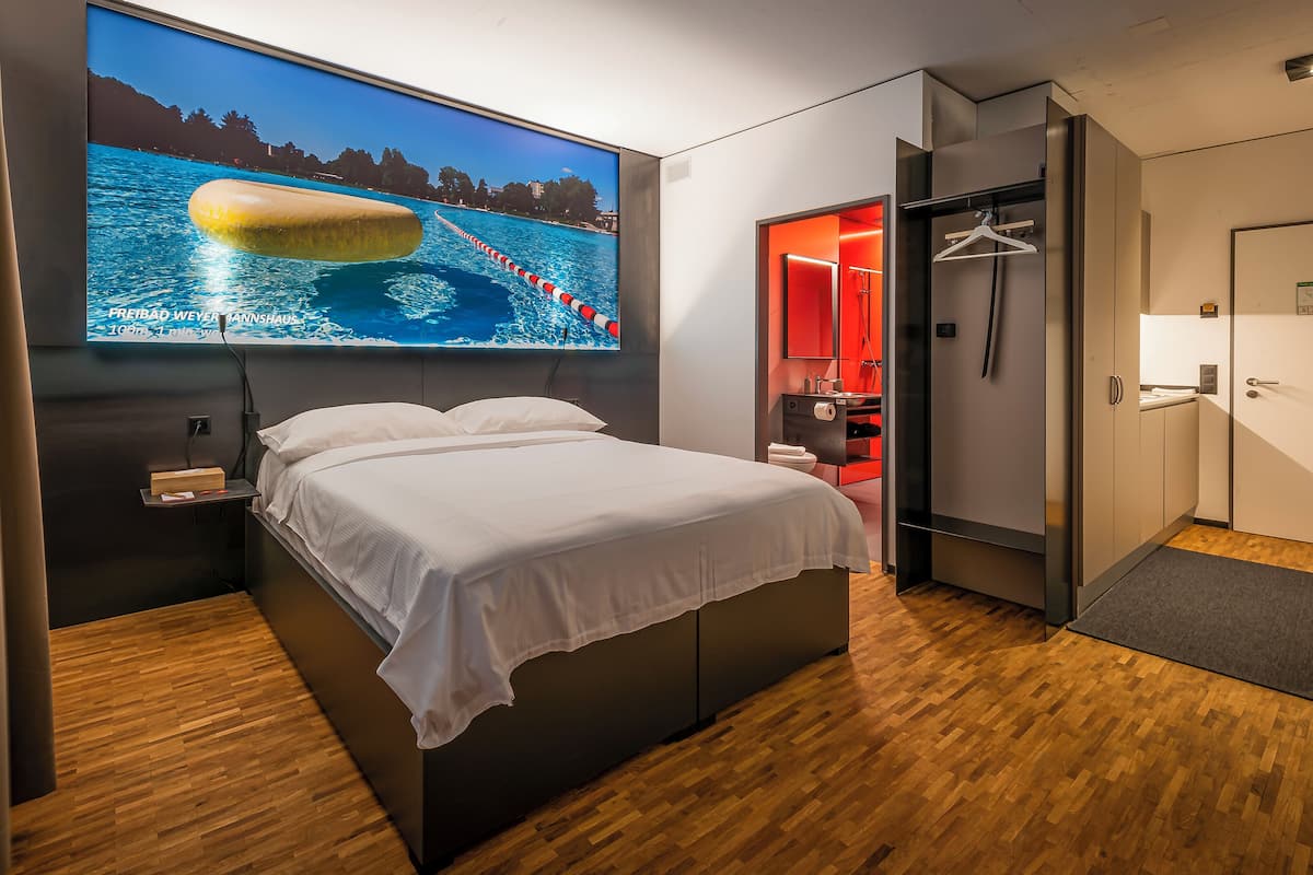 A modern hotel room with a large bed, a television displaying a swimming pool, a red bathroom, and a wooden floor.