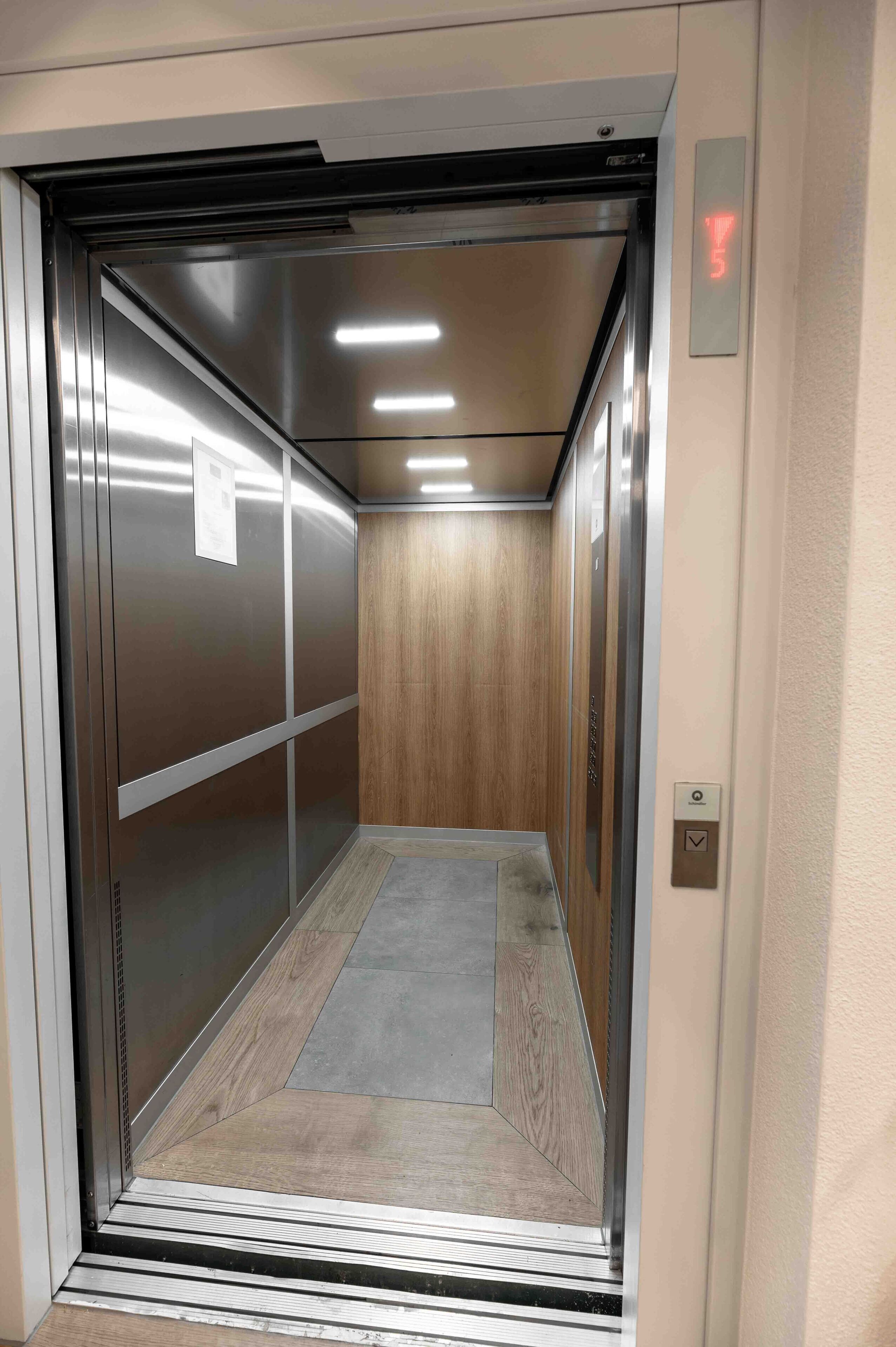 An elevator interior with a metallic finish, a wooden panel wall, and a tiled floor.