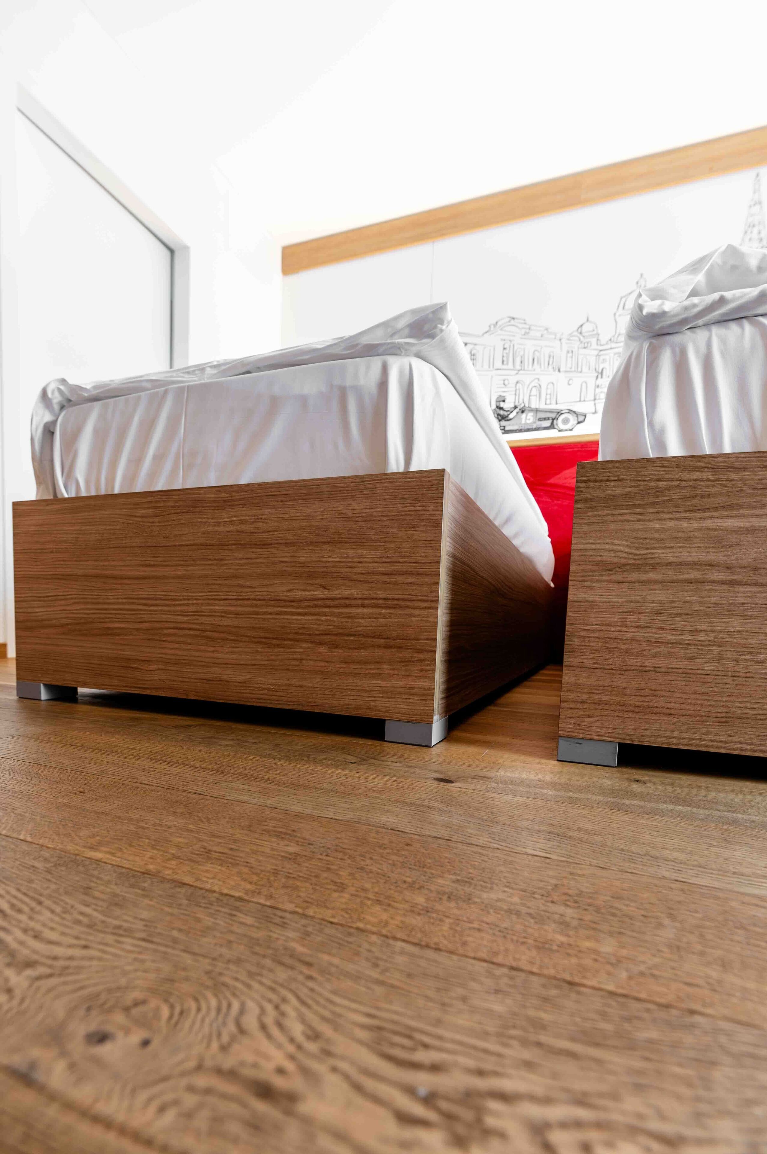A bed with a wooden frame and a red pillow, positioned in a room with a wooden floor and a white wall.