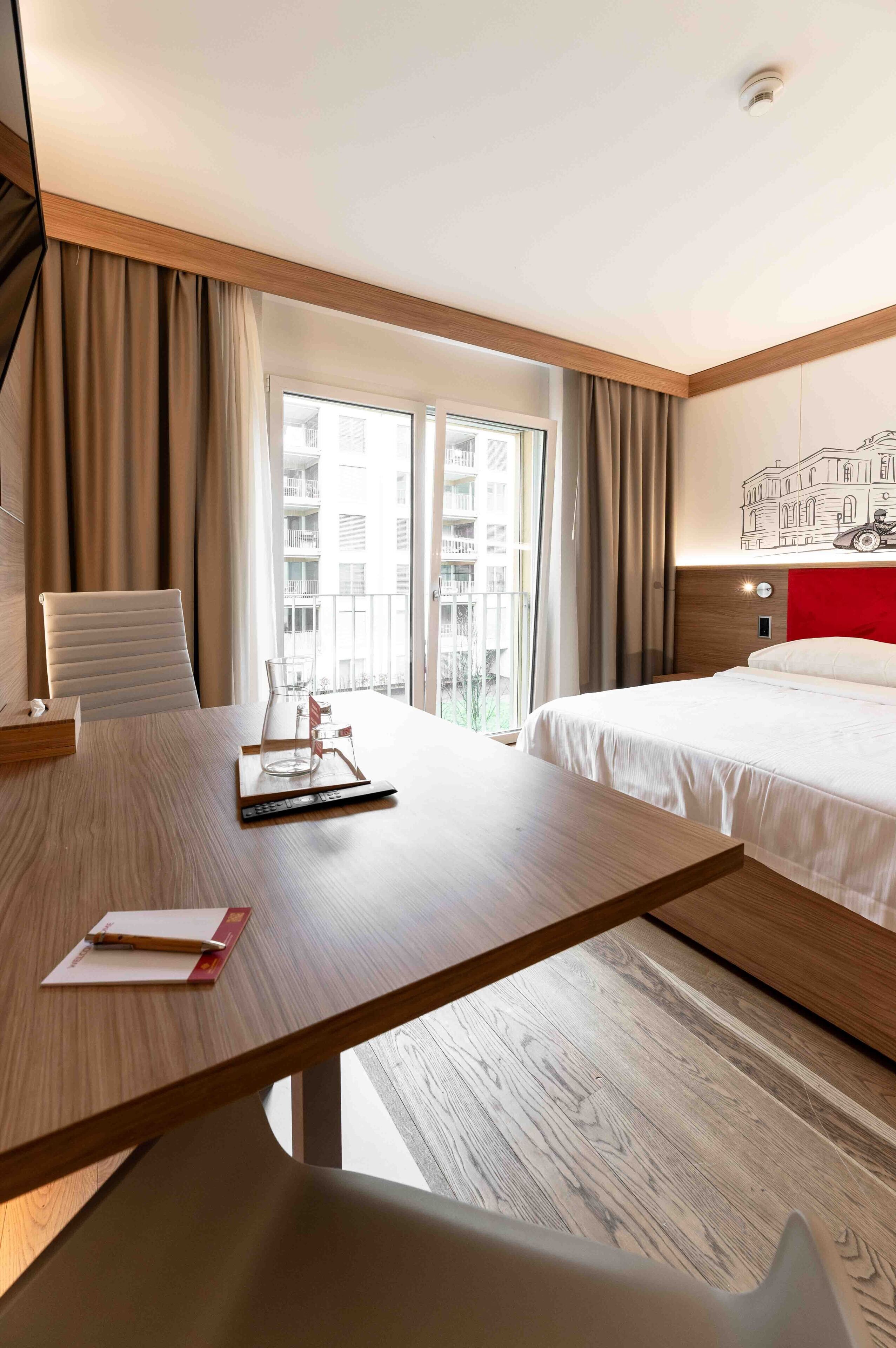 A hotel room with a wooden desk, a chair, a bed, and a view of the city through large windows.