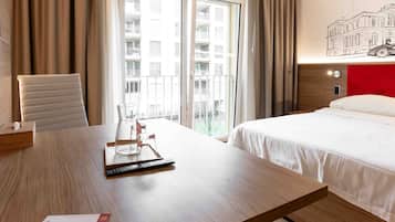 A hotel room with a wooden desk, a chair, a bed, and a view of the city through large windows.
