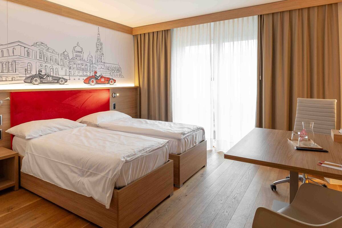 A hotel room with a wooden floor, a bed with a red headboard, a desk with a chair, and a large window with curtains.