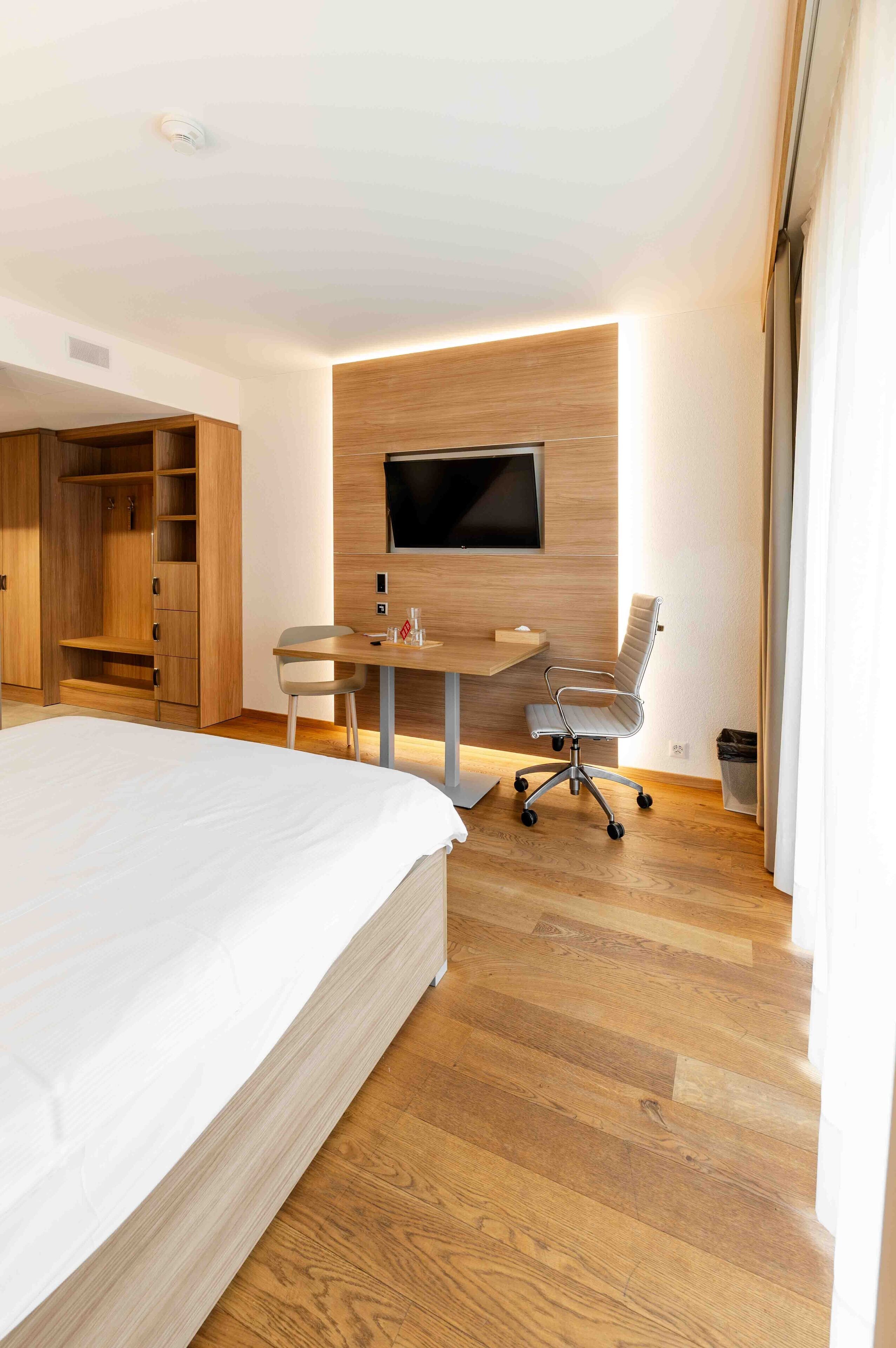 Habitación de hotel moderna con una cama grande, un escritorio con silla, un televisor montado en la pared y piso de madera.