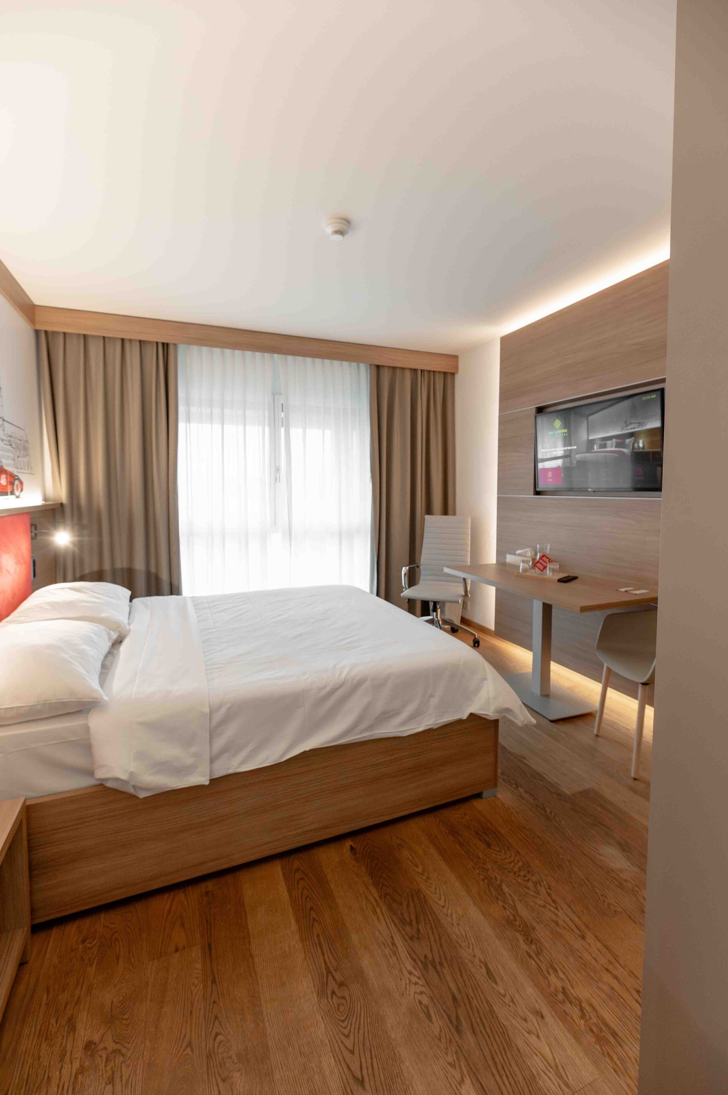 A modern hotel room with a wooden bed, a desk, and a TV on the wall.