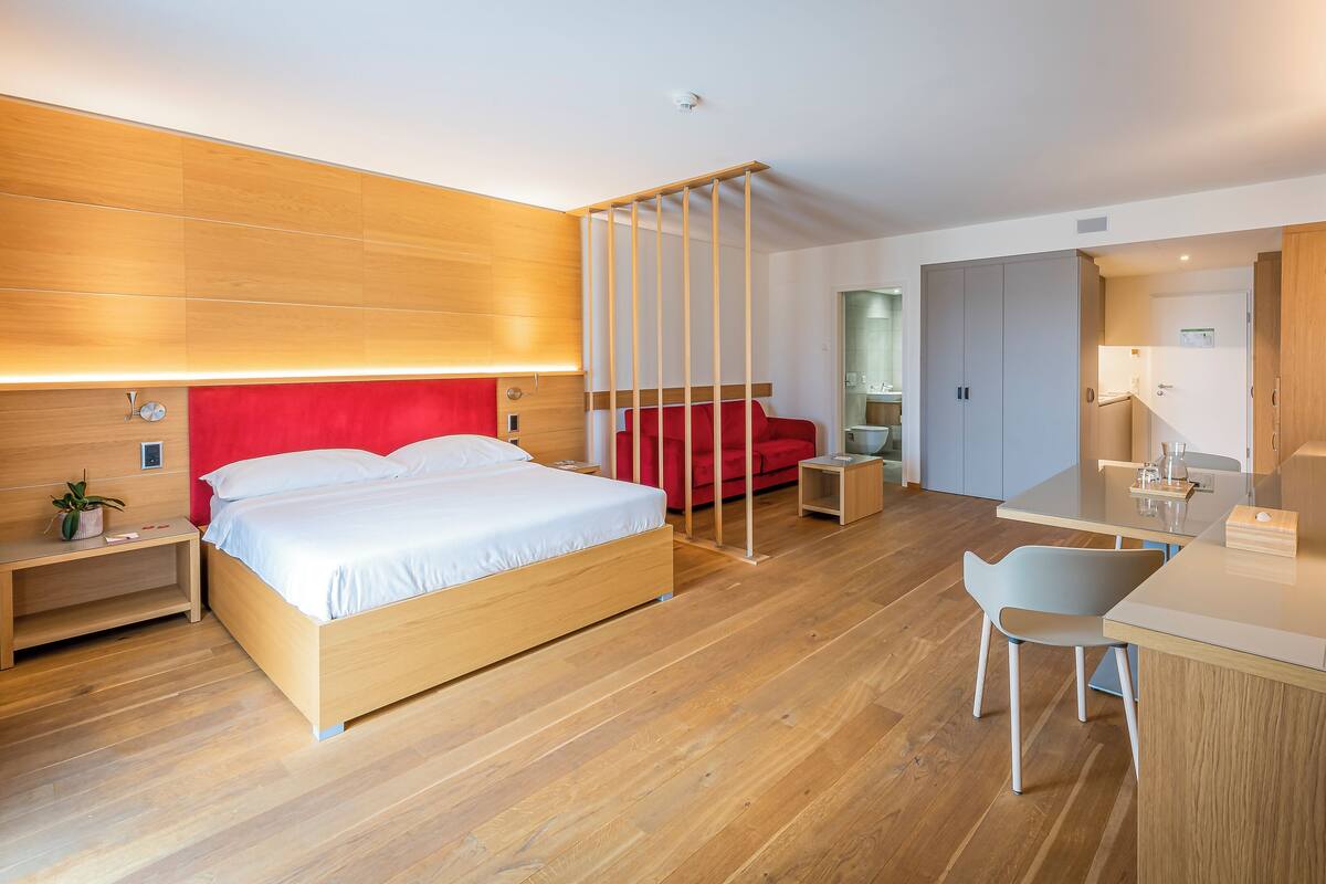 A modern hotel room with a wooden floor, a bed with white linens, a red sofa, a desk with a chair, and a bathroom visible in the background.