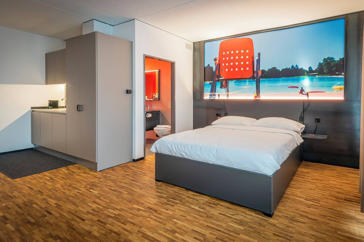 A modern hotel room with a bed, a desk, and a large window with a beach view.