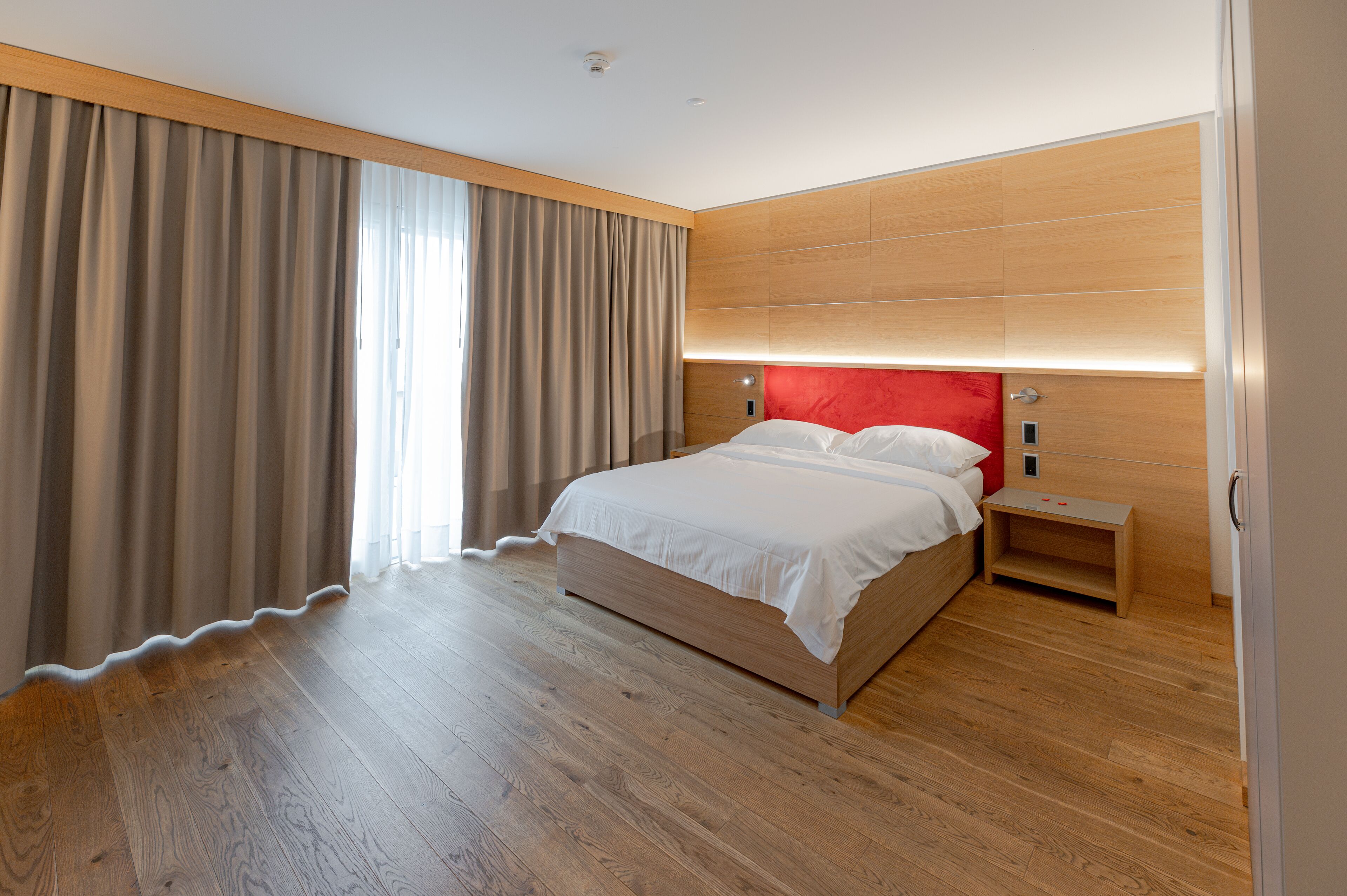 Een moderne hotelkamer met een houten vloer, een groot bed met witte beddengoed, een rood hoofdeinde en een nachtkastje.