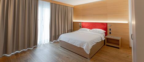 Una habitación de hotel moderna con piso de madera, una cama grande con ropa blanca, un cabecero rojo y una mesita de noche.