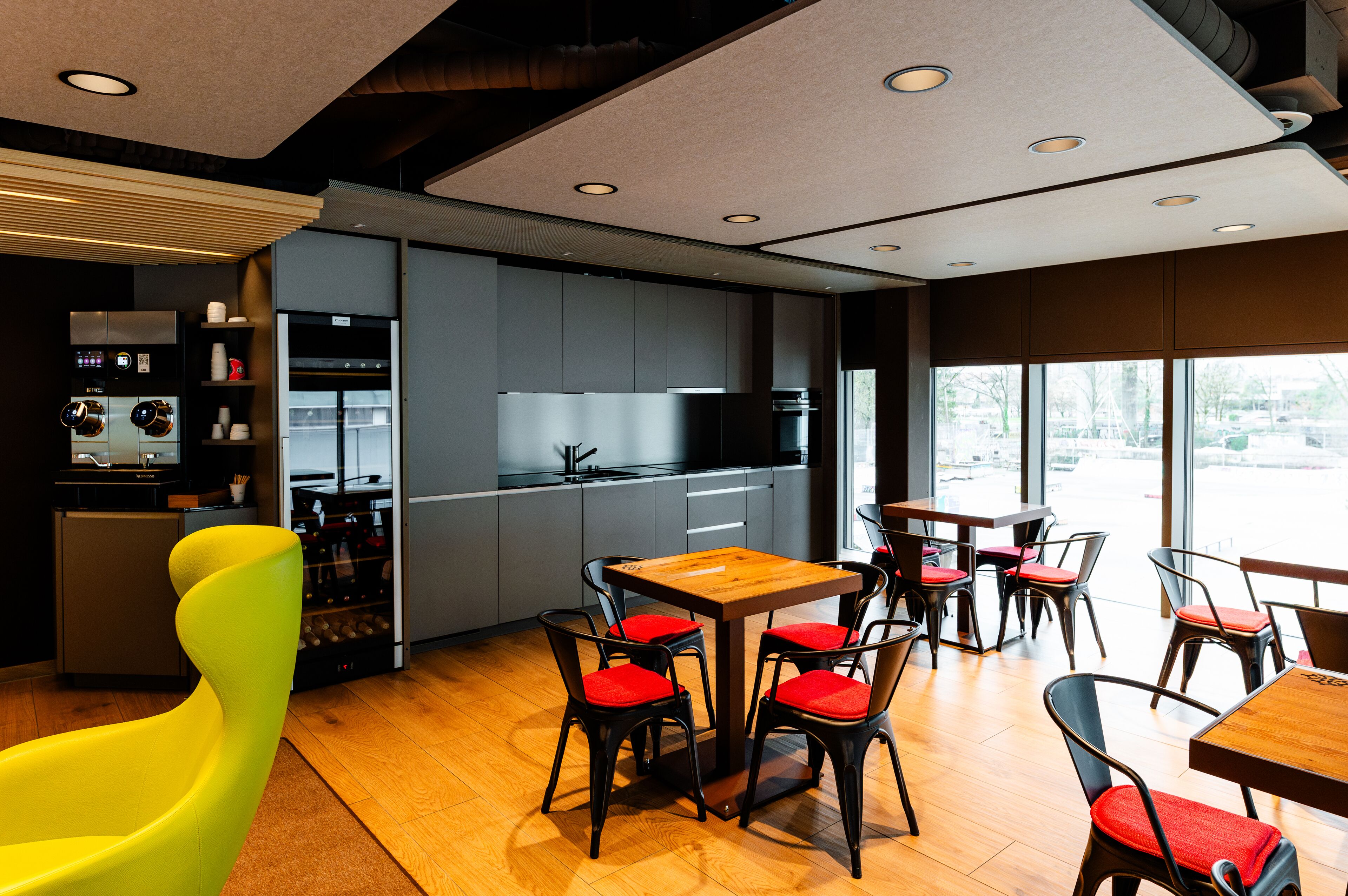 A modern kitchen with built-in appliances, a dining area with wooden tables and red chairs, and large windows offering a view.