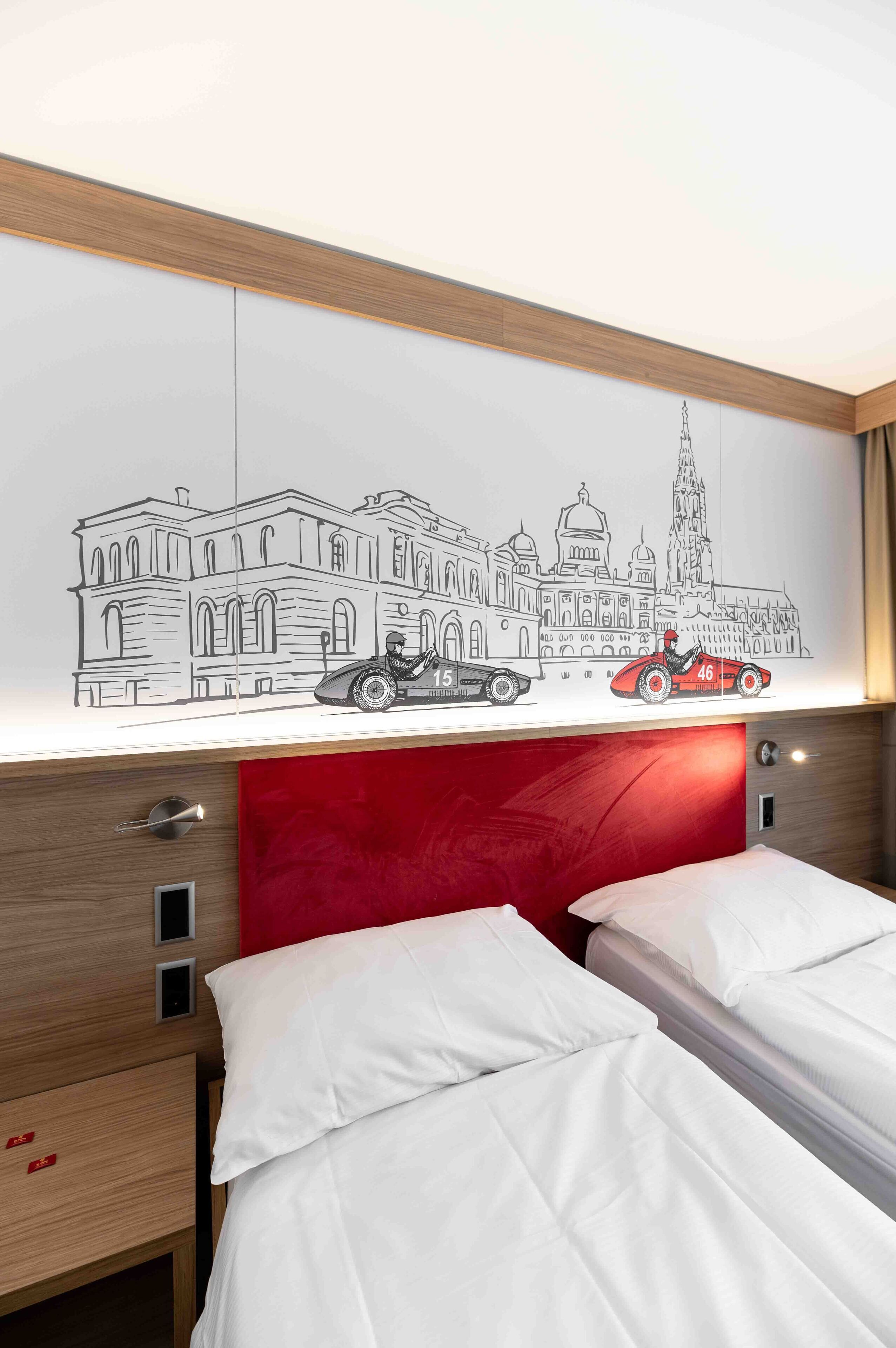 Hotellihuone, jossa on punainen sängynpääty, valkoisilla lakanoiden koristeltu sänky, seinälle ripustettu taideteos, jossa on kuvattuna kaupunkimaisema vanhoine autoineen, sekä puinen yöpöytä, jossa on yksi laatikko.