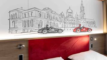 A hotel room with a red headboard, a bed with white bedding, a wall-mounted artwork depicting a cityscape with vintage cars, and a wooden nightstand with a drawer.