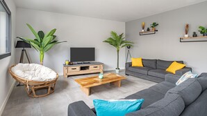 A modern living room with a grey sofa, a wooden coffee table, a wicker chair, and potted plants.