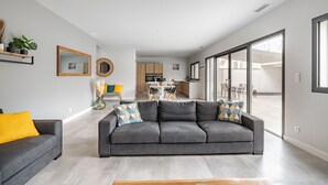 A modern living room with a grey sofa, a smaller grey armchair, and a coffee table. There is a kitchen area in the background with a dining table and chairs.