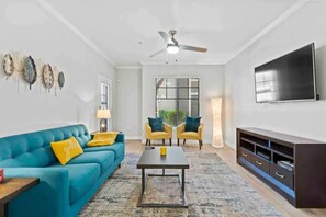 A living room with a teal sofa, two yellow armchairs, a coffee table, a TV, and a ceiling fan.