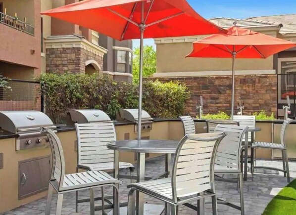 An outdoor patio with metal chairs and tables, red umbrellas, and a barbecue grill.