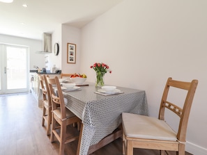 A dining area with a table set for four, wooden chairs, a vase with red flowers, and a kitchen in the background.