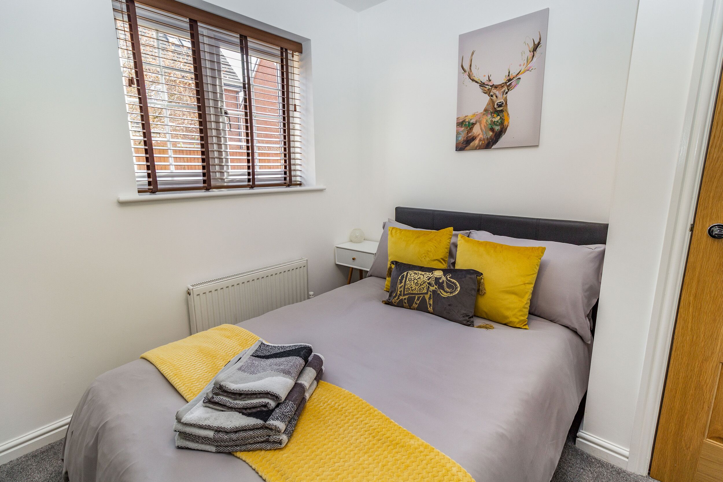 Una cama bien hecha con cubrecama gris y amarillo, una manta gris y blanca doblada, y una manta amarilla. Se ve una ventana con persianas, un radiador blanco y un cuadro colgado en la pared con un ciervo.