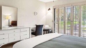 Ein Schlafzimmer mit Bett, Schreibtisch, Stuhl, Spiegel, Uhr und Fenster mit Jalousien.