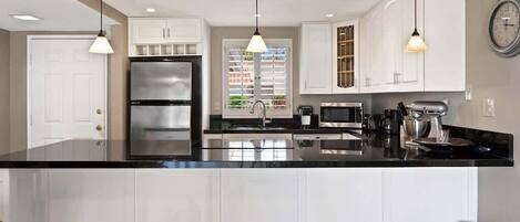 Une cuisine moderne avec des meubles blancs, un plan de travail noir, des appareils en acier inoxydable, des luminaires suspendus et une horloge visible au mur.