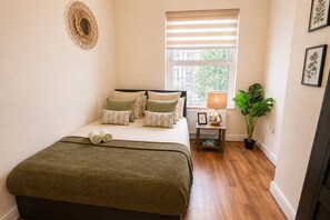 A bedroom with a bed, pillows, a bedside table with a lamp, a plant, and a window with blinds.