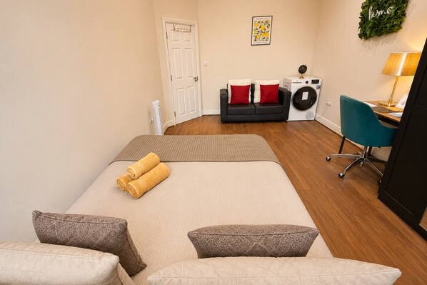 A hotel room with a bed, two pillows, a desk with a chair, a washing machine, and a wall-mounted lamp.