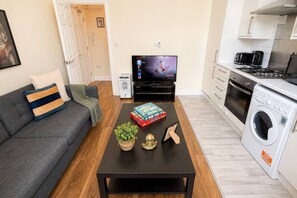 A modern living room with a sofa, a coffee table with books and a plant, a TV, and a kitchen area with a washing machine.