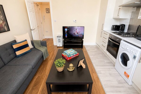 A modern living room with a sofa, a coffee table with books and a plant, a TV, and a kitchen area with a washing machine.