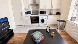 Een moderne keuken met witte kasten, een magnetron, oven en een wasmachine. Er staat een zwarte tafel met gezelschapsspellen en een klein gouden beeldje.