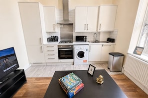 A modern kitchen with white cabinets, a microwave, oven, and a washing machine. There's a black table with board games and a small golden statue. - Cozy 2-bedroom Apt in Shepherds near Westfield (London)