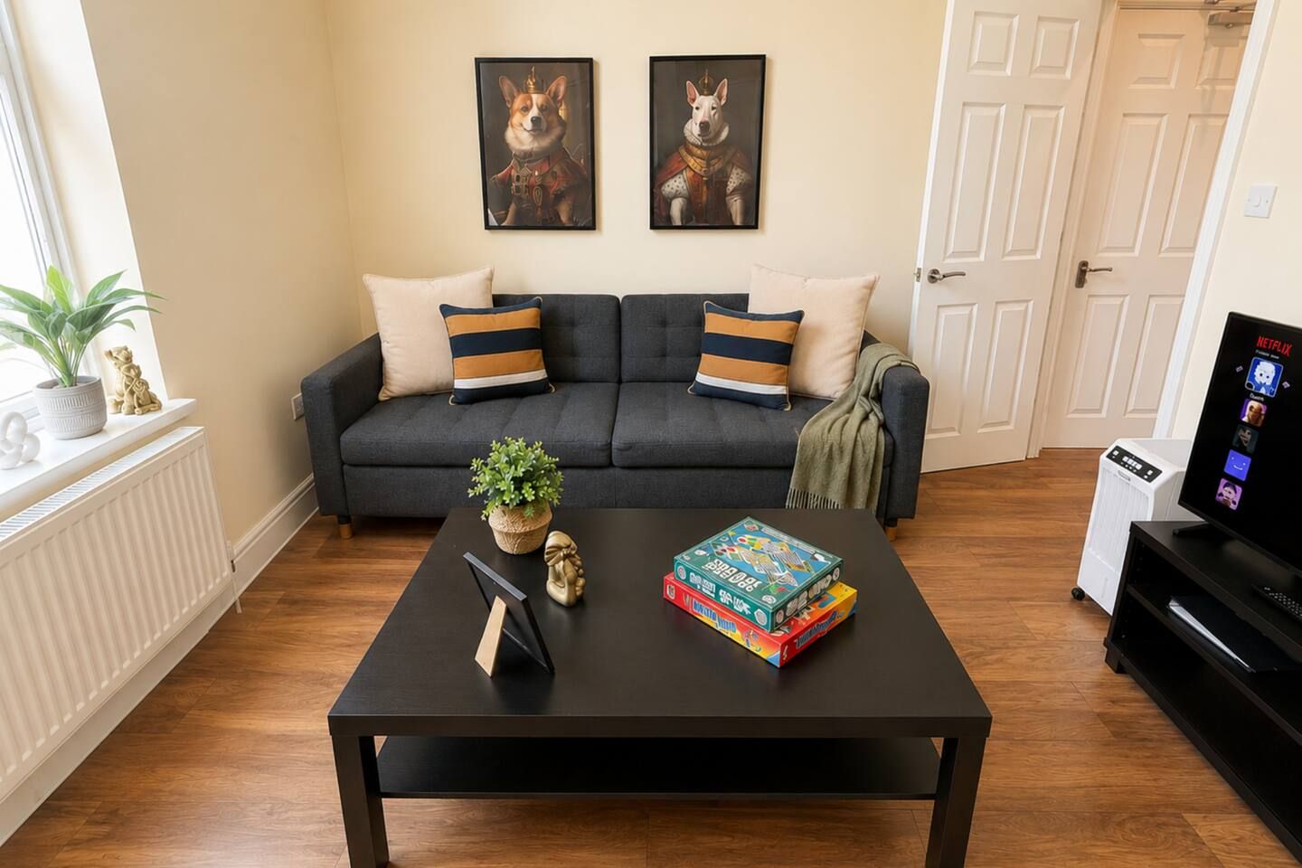 A living room with a black sofa, two framed pictures on the wall, a coffee table with books and a plant, and a TV on a stand.
